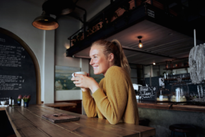 Kvinna i gul tröja som håller i en kaffekopp och ler vid ett träbord på ett mysigt kafé med rustik inredning, perfekt för dig som studerar på distans.