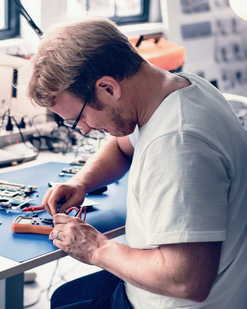 En man med glasögon som reparerar en elektronisk enhet vid en arbetsbänk med verktyg och kablar i en ljus arbetsmiljö, vilket visar på den precision som kännetecknar en skicklig mjukvaruutvecklare i arbete.