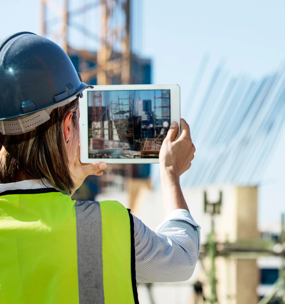 En byggnadsingenjör, klädd i en hjälm och skyddsväst, använder en surfplatta för att fånga en bild på en livlig byggarbetsplats.