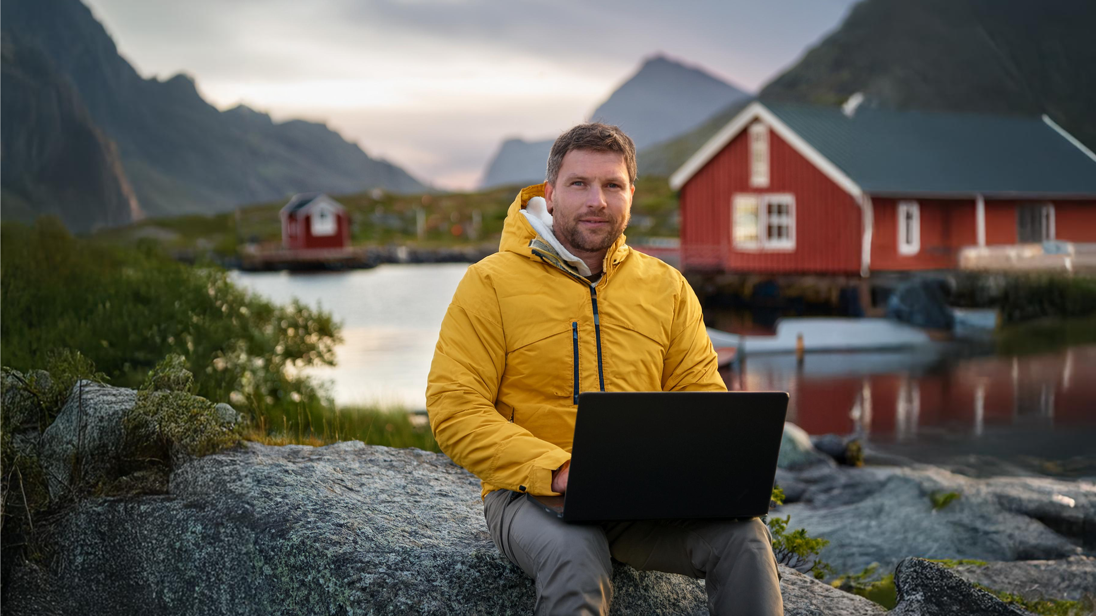 Affärsutvecklare njuter av lugnet i naturen och arbetar på sin bärbara dator utomhus vid en fridfull sjö, omgiven av pittoreska röda hus och majestätiska berg i bakgrunden.