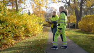 Två kvinnor i hi-vis-kläder, en en arbetsledare från hållbar grönyteförvaltning i Ängelholm, diskuterar nära en stig omgiven av höstlöv och träd i parken.