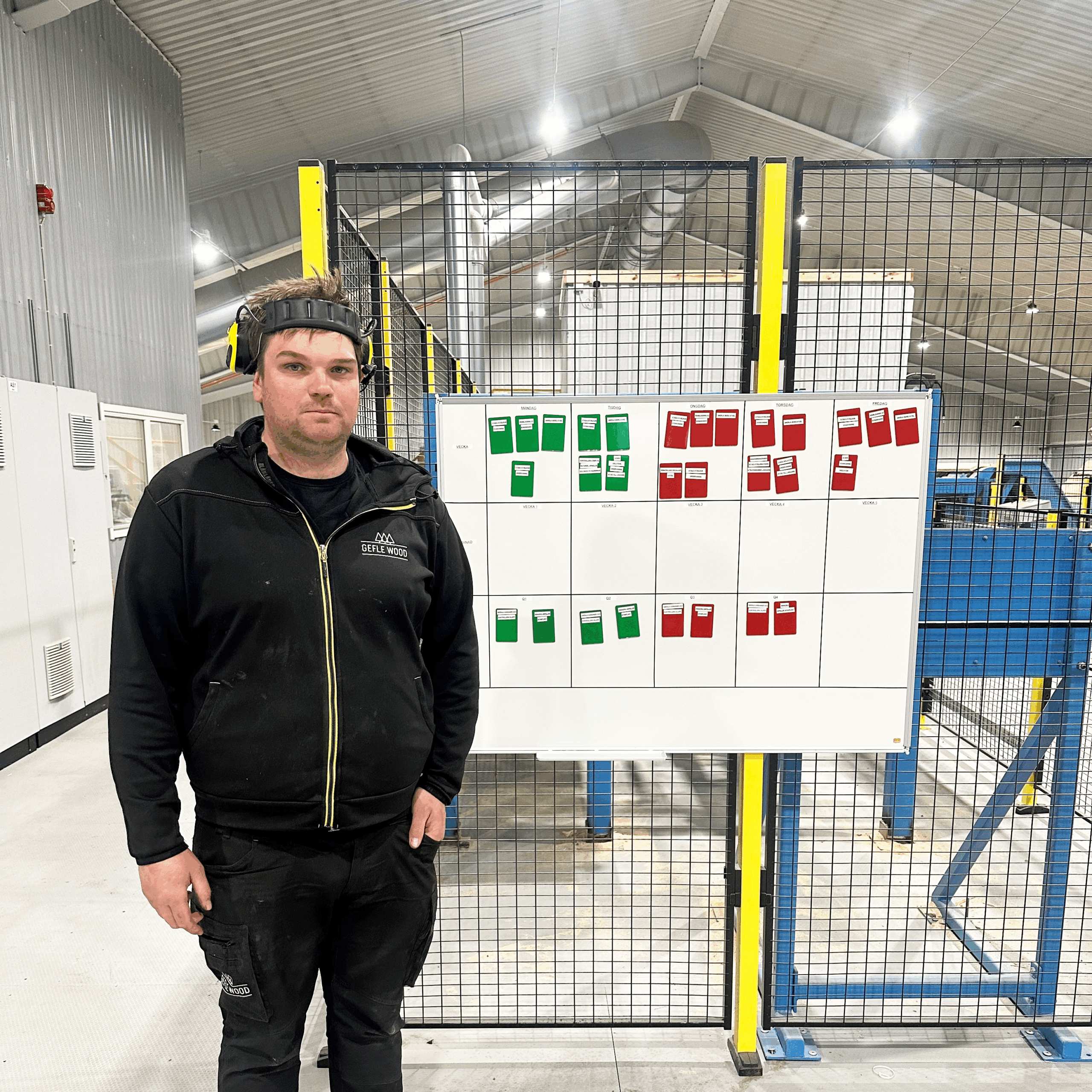 En man står inomhus vid en whiteboardtavla med röda och gröna kort i ett rutnät och markerar Produktionsutveckling i en industriell miljö.