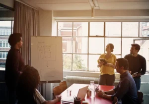 Fyra personer i ett mötesrum diskuterar annonsering i sociala medier, med hänvisning till ett diagram på en whiteboardtavla vid fönstret, med bärbara datorer på bordet.