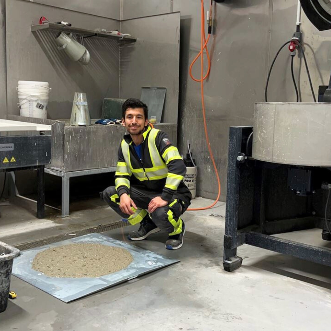 En betongingenjör, klädd i högviskväxel, knäböjer bredvid en våt betongblandning i en industriell miljö.
