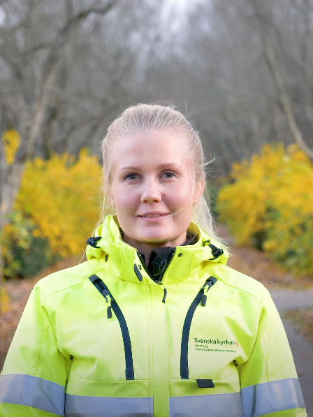 En person som bär en knallgul jacka står på en gångväg, omgiven av gula blommor och kala träd, förkroppsligar andan hos en arbetsledare i hållbar grönyteförvaltning i Ängelholm.