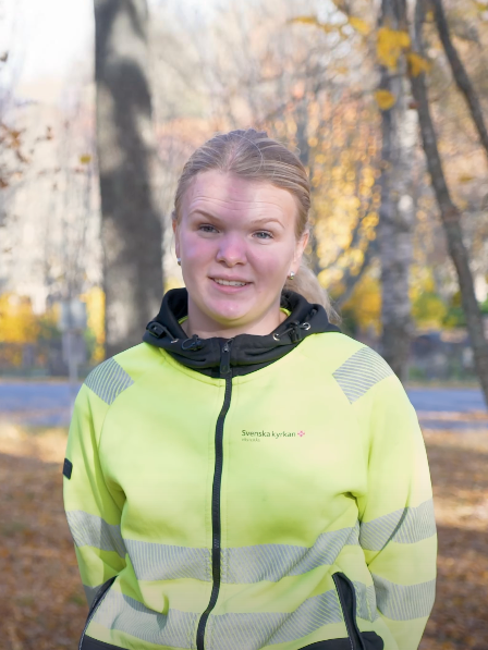 En person i neongul jacka står ute en solig höstdag, med träd och nedfallna löv runt omkring, förkroppsligar rollen som arbetsledare inom hållbar grönyteförvaltning.