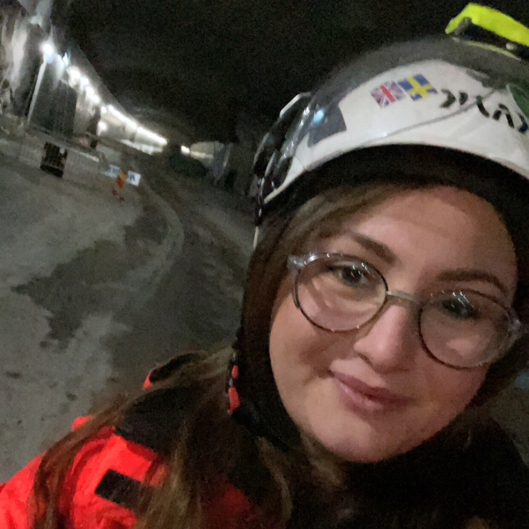 Betongingenjör i skyddsutrustning och hjälm tar en selfie inne på en svagt upplyst tunnelbyggarbetsplats.