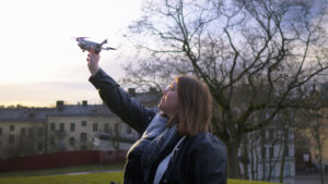 En fjärrpilot håller upp en liten drönare i luften utomhus nära träd och byggnader en molnig dag.