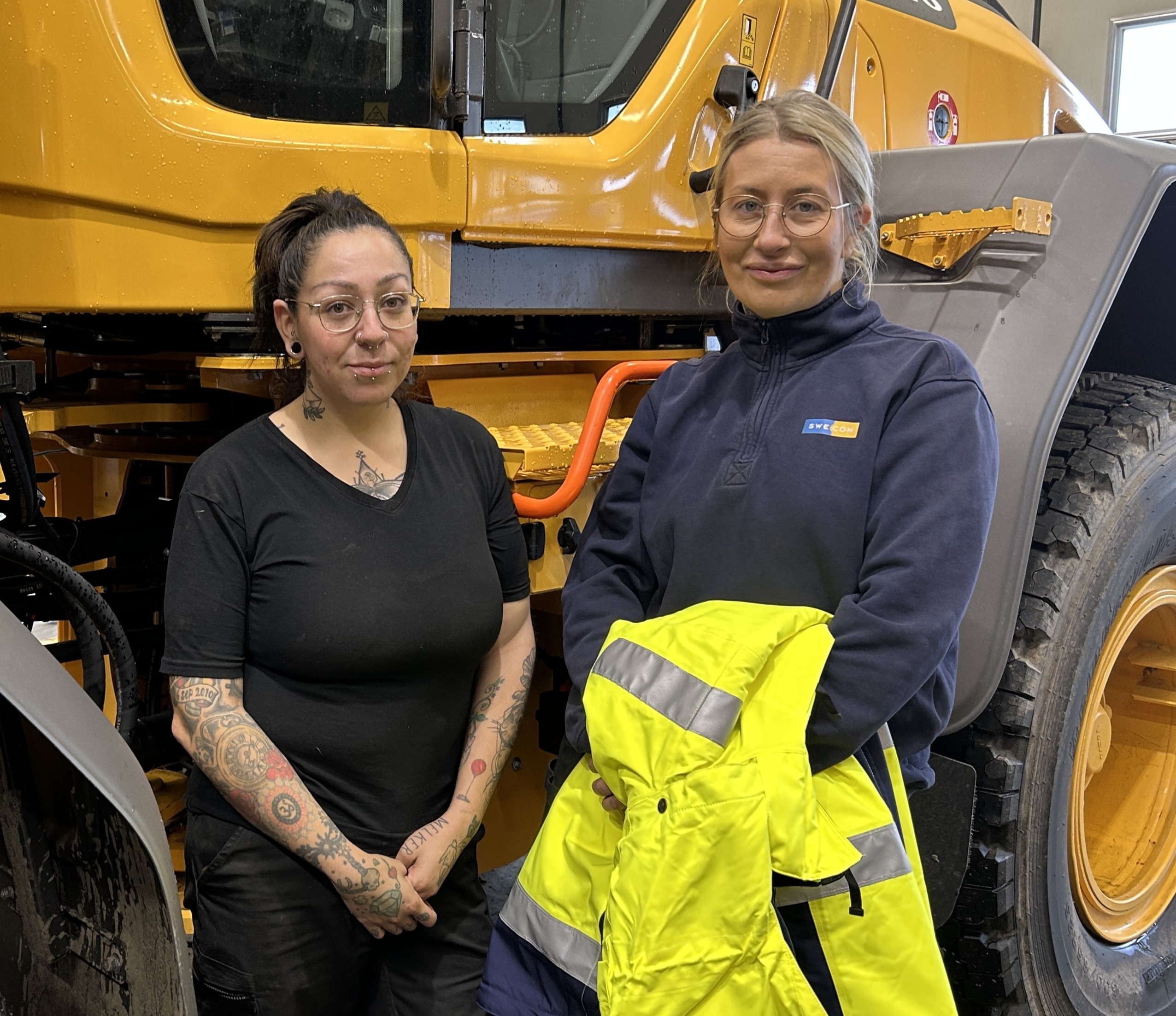 Två kvinnor står framför en stor gul byggbil, den ena med en varselväst - redo för arbete som tekniker tunga maskiner och fordon.