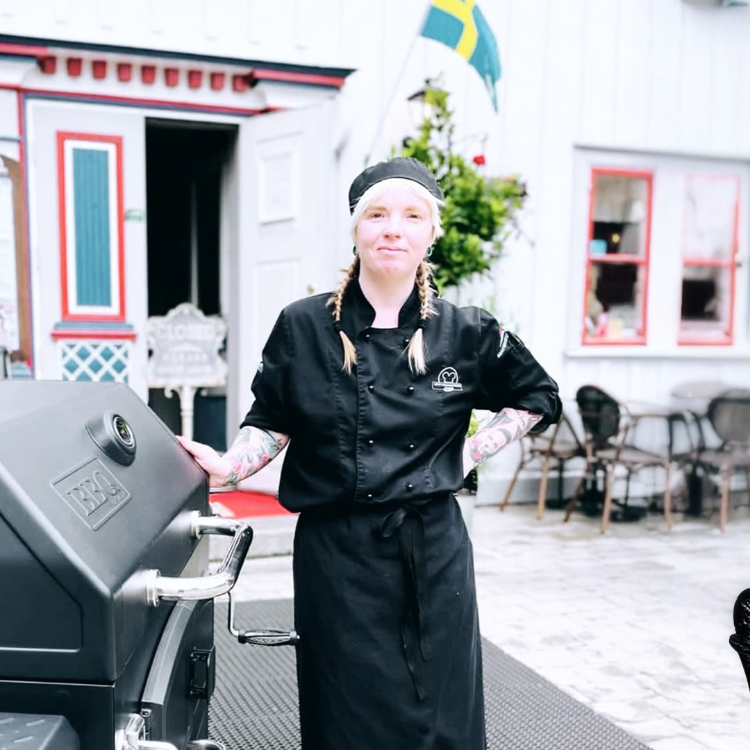 En kock i svart uniform står vid en grill utanför en restaurang och representerar stolt Gastronomiledare - Framtidens mat och protein, med en svensk flagga som vajar i bakgrunden.