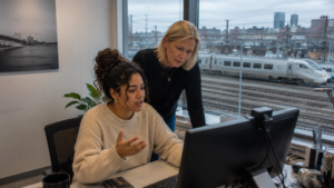 Två kvinnor, varav en elingenjör, arbetar vid en dator på ett kontor med utsikt över tågspår och ett passerande tåg.