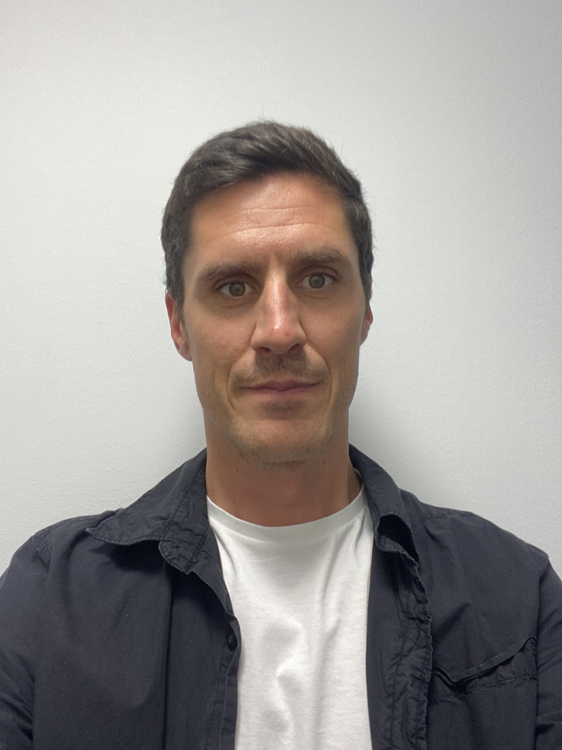 Man med kort mörkt hår, en Elingenjör, i vit t-shirt och svart skjorta står mot en enfärgad ljus vägg.