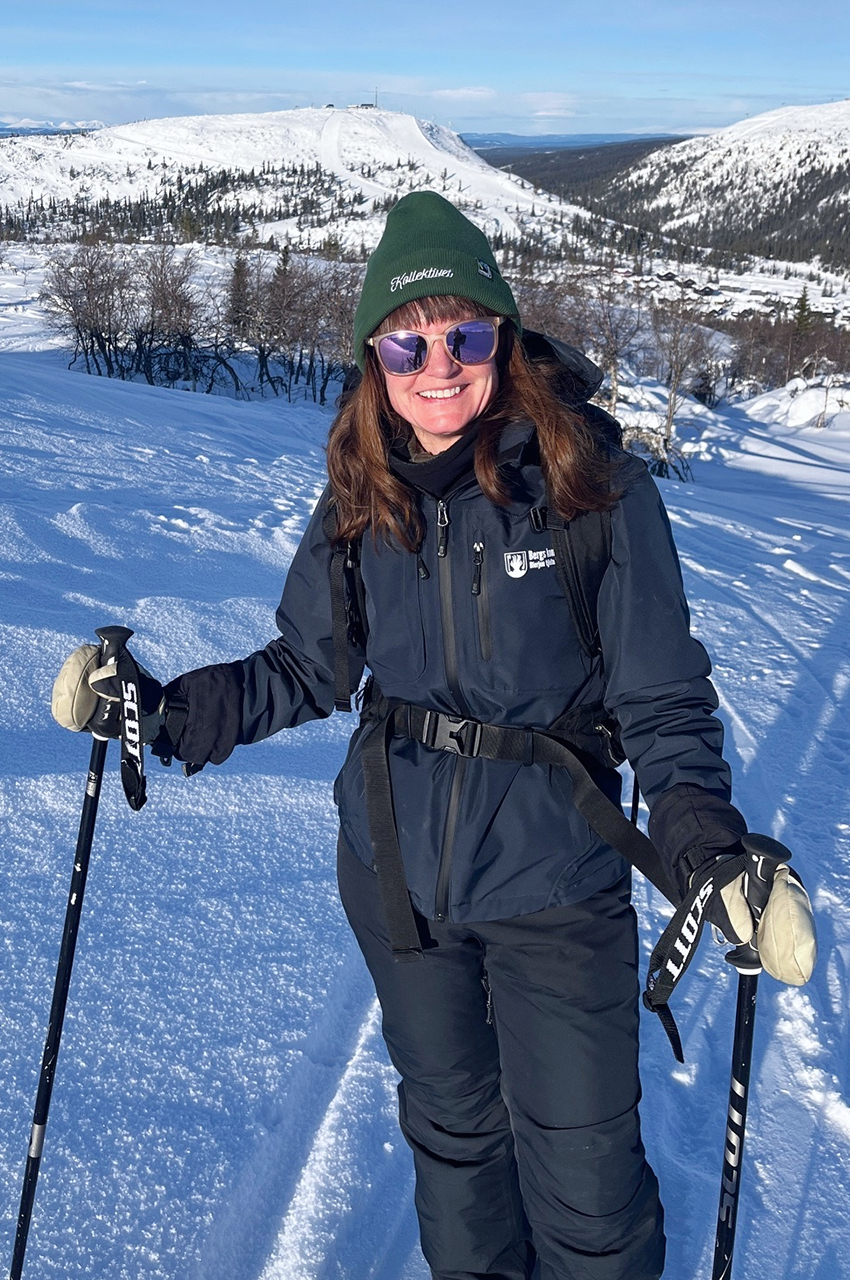 Person som ler medan du åker skidor på ett snöigt berg, bär en mörk outfit och solglasögon. Som en affärsutvecklare som navigerar i besöksnäringen njuter de av spänningen i det ljusa, soliga landskapet i bakgrunden.