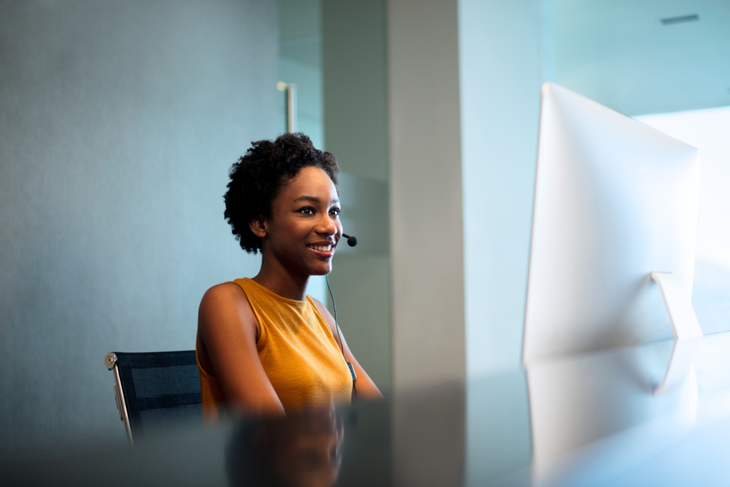 Här är den reviderade alt-texten: En person som bär ett headset och ler, sitter vid ett skrivbord med en stor datorskärm i en kontorsmiljö och visar upp sin senaste utbildning vid YH Akademin under sin karriärbyte utbildning.