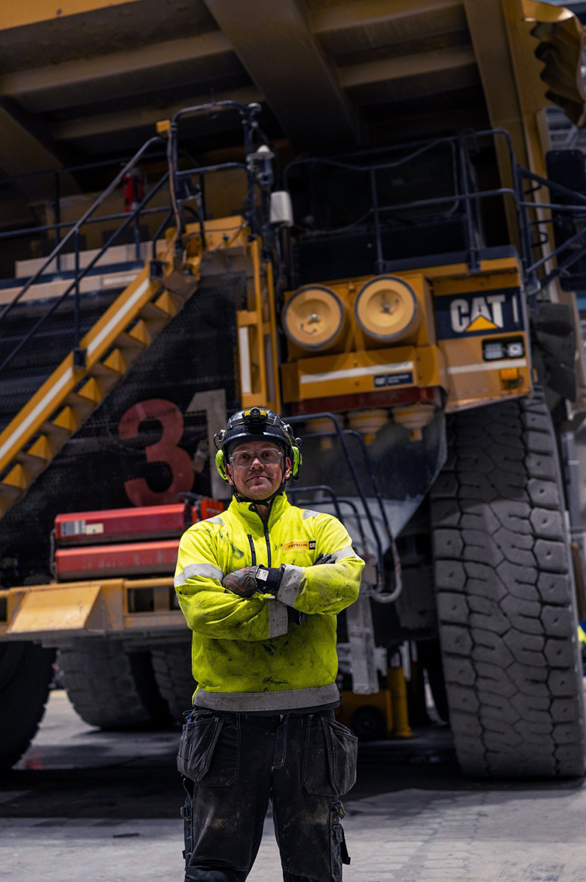 En Tekniker i gul jacka står självsäkert framför en stor industriell gruvlastbil inne i en anläggning och visar upp expertis inom tunga maskiner och fordon.