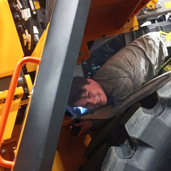 En person som ler medan han inspekterar eller reparerar en stor gul traktor nära dess hjul, vilket exemplifierar expertisen hos en Tekniker tunga maskiner och fordon.