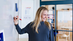 En leende socialpedagog håller i en röd tuschpenna när hon står vid en whiteboard i ett ljust klassrum.