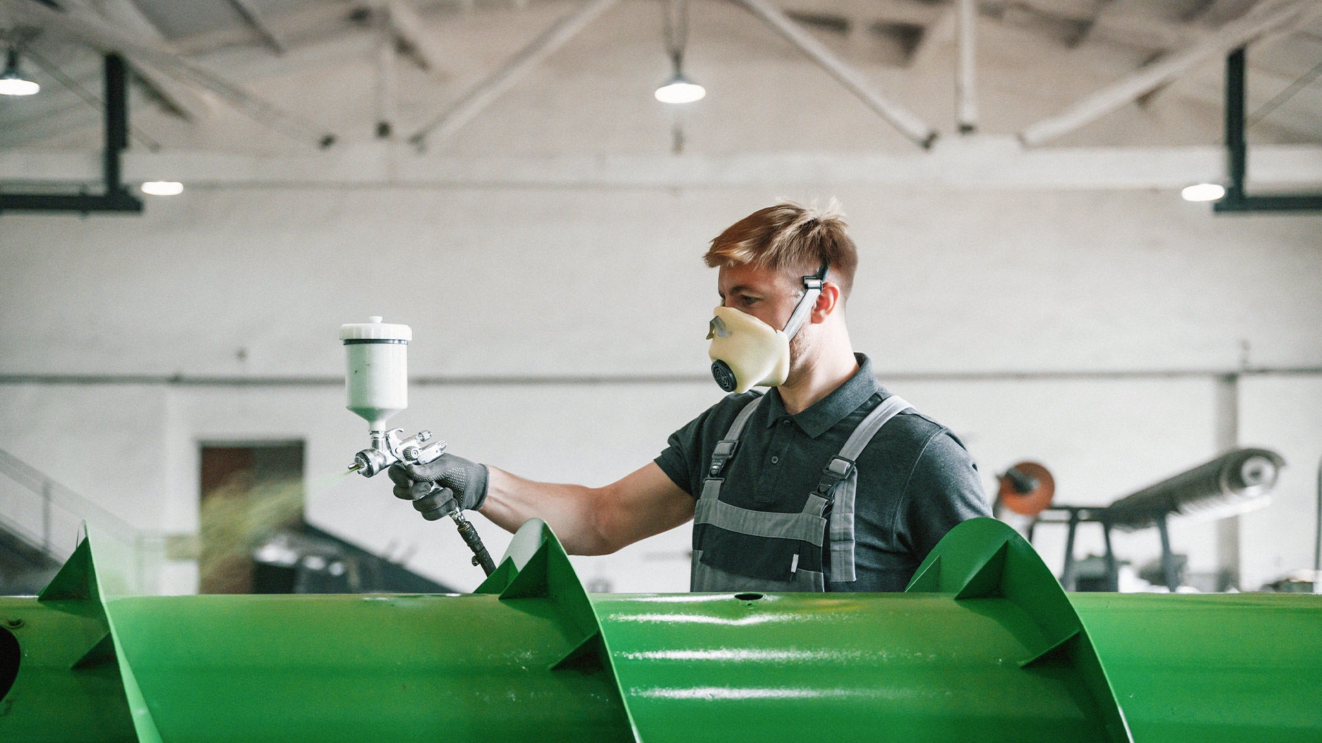 En Ytbehandlingsspecialist iförd mask och handskar spraymålar ett stort grönt metallföremål i en industrimiljö.