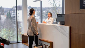 Två kvinnor interagerar i en hotellreception; den ena, möjligen hotellchefen YH-flex, ler varmt när den andra checkar in med sin resväska.