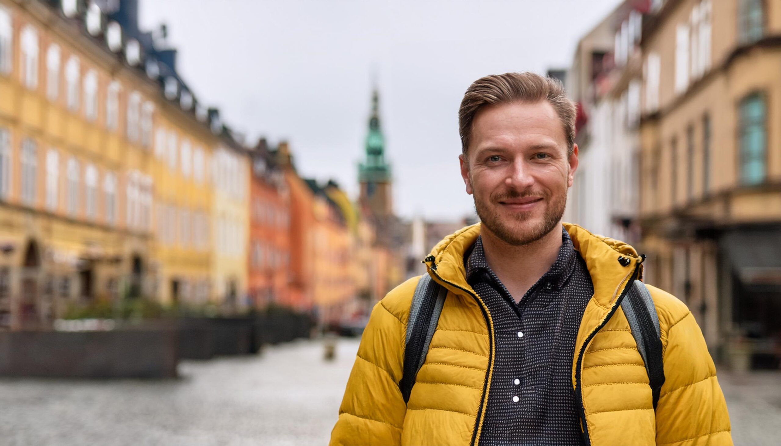 En man i gul jacka står och ler på en kullerstensgata kantad av färgglada byggnader och tar en paus från sin tjänstledighet för studier.