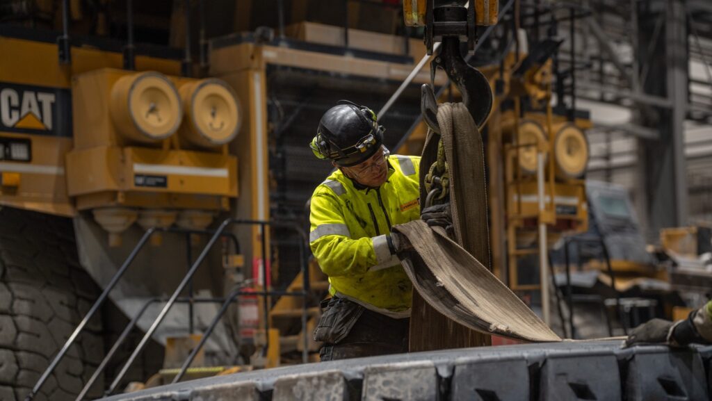 Arbetare i säkerhetsutrustning använder tunga maskiner i en industriell miljö med stora däck i bakgrunden.