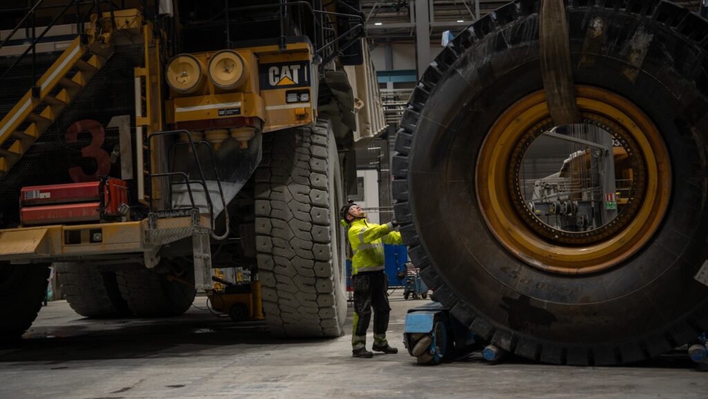 Arbetare i gul jacka inspekterar ett gigantiskt däck i en industriell lagermiljö.