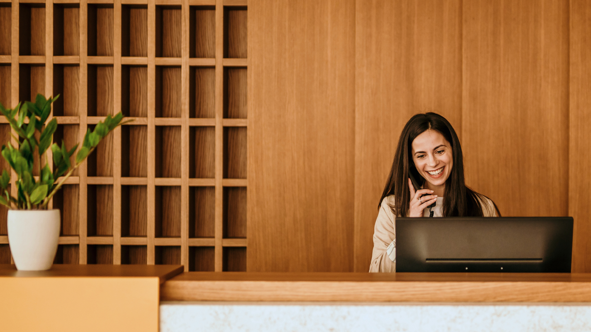 En leende receptionist pratar i telefon bakom ett skrivbord i ett modernt träkontor, med en krukväxt på bänken, vilket exemplifierar den välkomnande atmosfären i ett yrke inom turism, hotell och restaurang