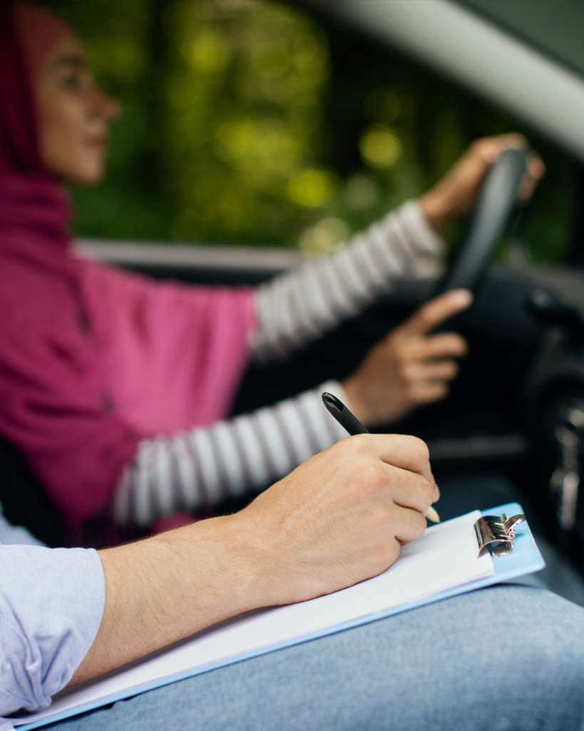 En person kör medan en annan, möjligen en instruktör, gör anteckningar på ett skrivunderlägg i passagerarsätet, vilket tyder på ett körprov eller ”Uppdragsutbildning för företag – Trafiklärare”.