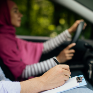 En kvinna i hijab kör bil, medan en instruktör – som deltar i programmet ”Uppdragsutbildning för företag – Trafiklärare” – gör anteckningar på ett skrivunderlägg från passagerarsätet.