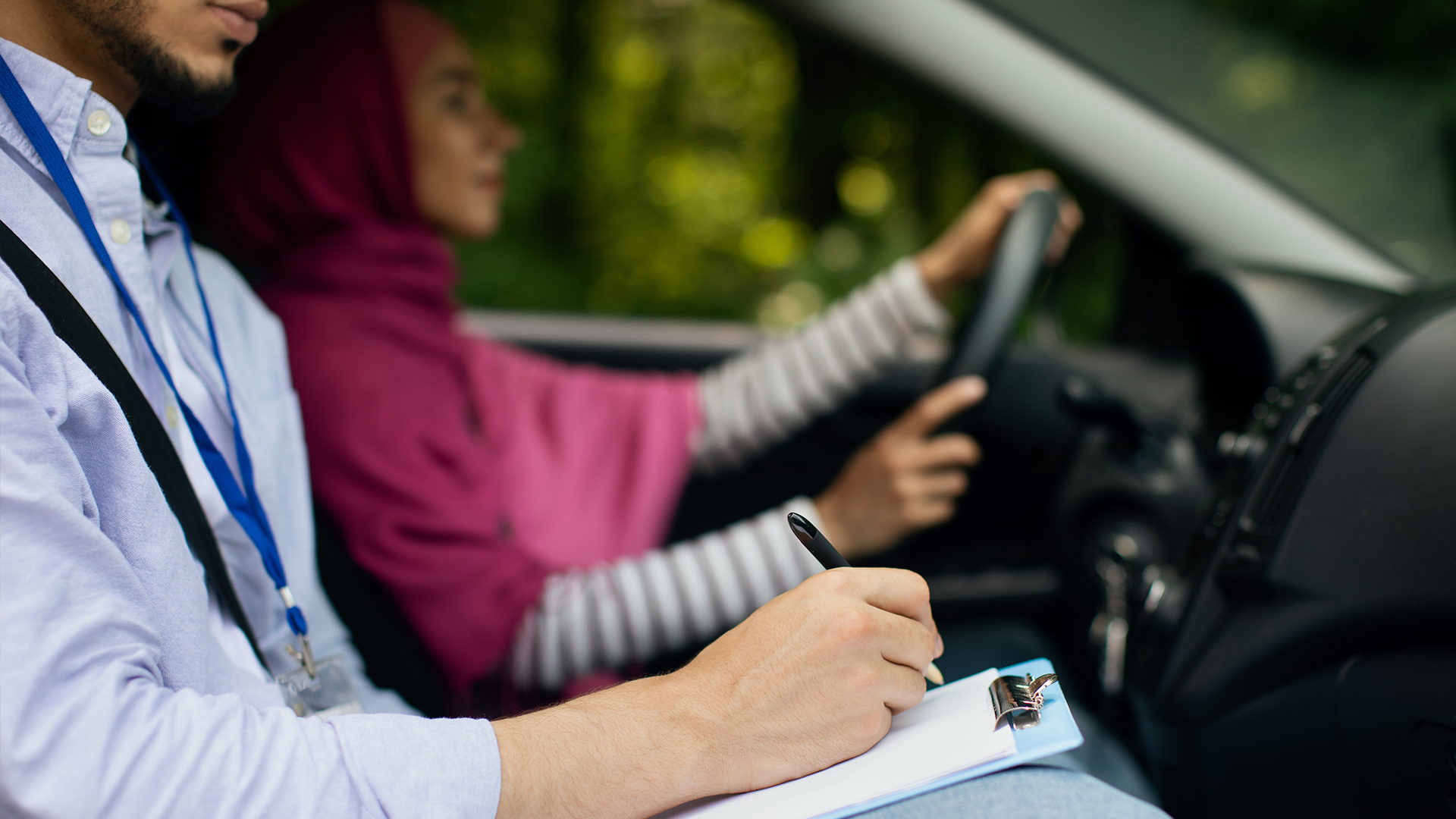 En kvinna i hijab kör bil, medan en instruktör – som deltar i programmet ”Uppdragsutbildning för företag – Trafiklärare” – gör anteckningar på ett skrivunderlägg från passagerarsätet.
