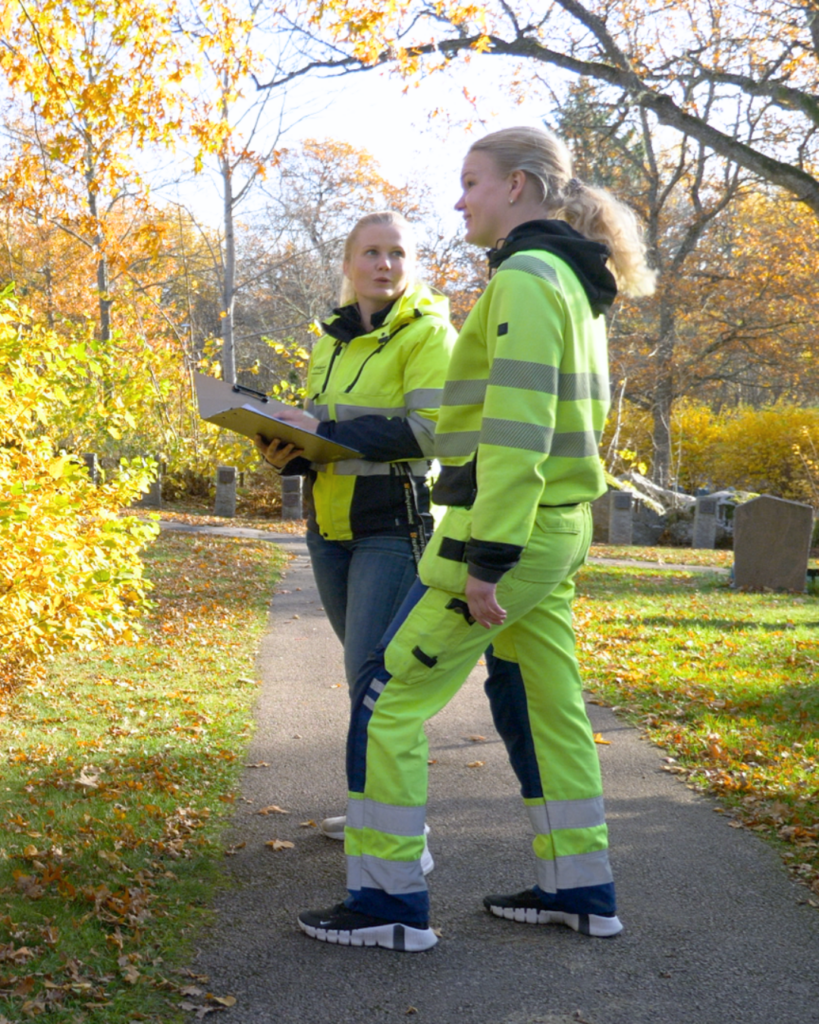 Två personer i synliga kläder, varav en ser ut att vara en arbetsledare utemiljö, samtalar på en solig parkstig omgiven av höstlöv.