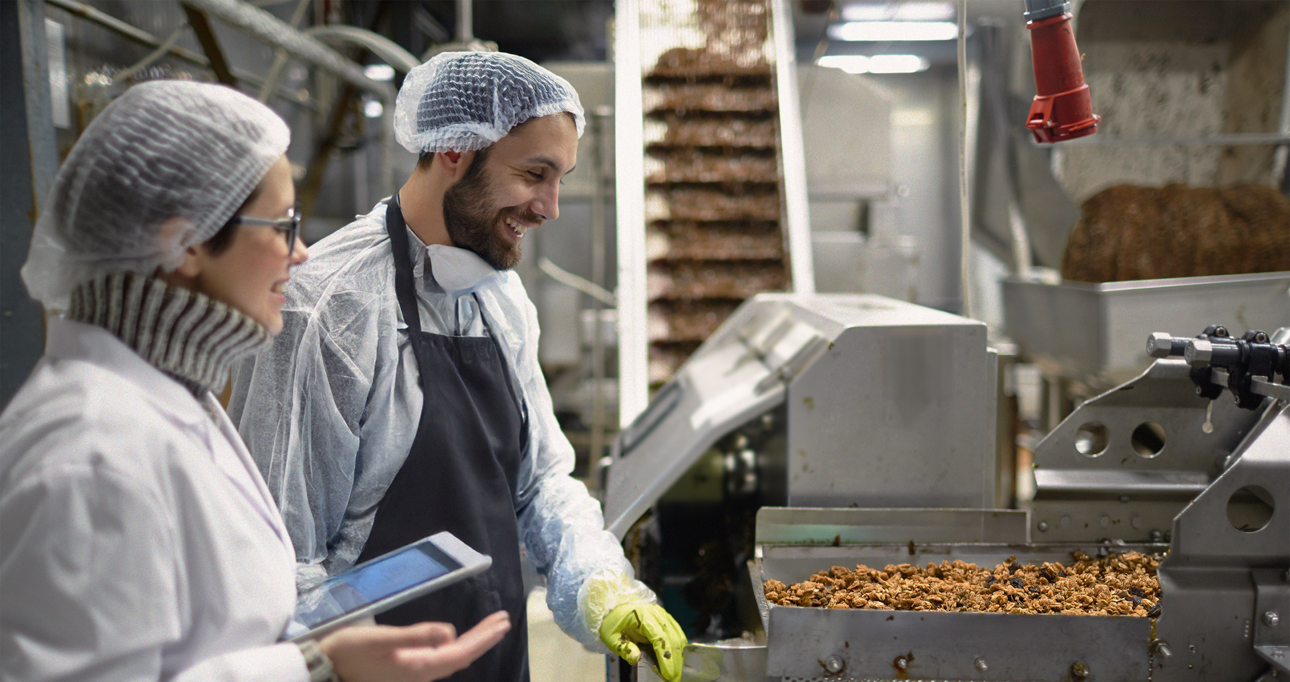 Två arbetare i skyddsutrustning, inklusive en produktionsansvarig livsmedelsutveckling, inspekterar maskiner för livsmedelsbearbetning och diskuterar produktionen i en fabriksmiljö.