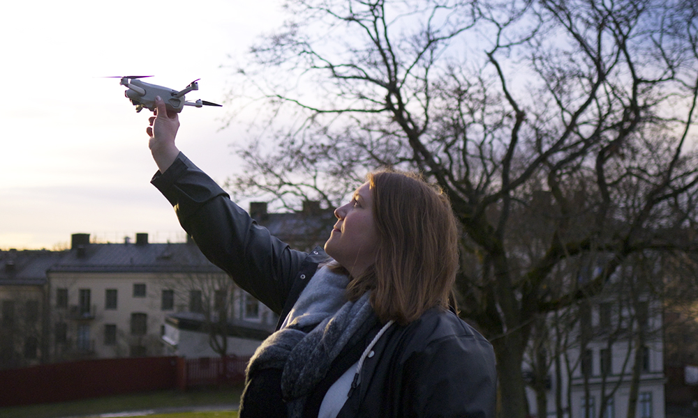En drönarpilot, placerad bland träden och byggnaderna, håller upp en drönare utomhus med övat självförtroende.
