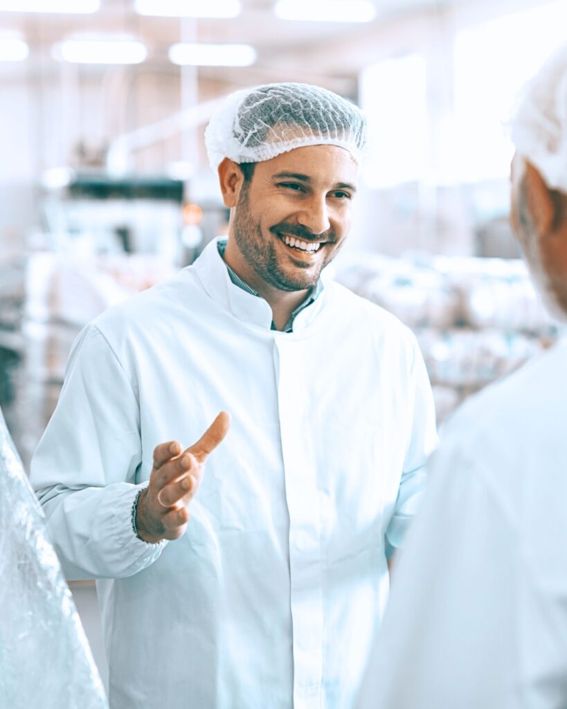 En livsmedelsoperatör i labbrock och hårnät ler varmt medan han chattar med en kollega i en starkt upplyst industrimiljö.