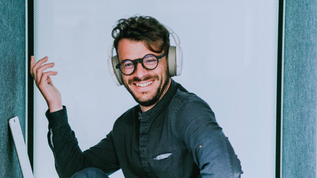 En leende person, som tar på sig runda glasögon och hörlurar, sitter avslappnad och påminner om en student som njuter av en paus på yrkeshögskola.