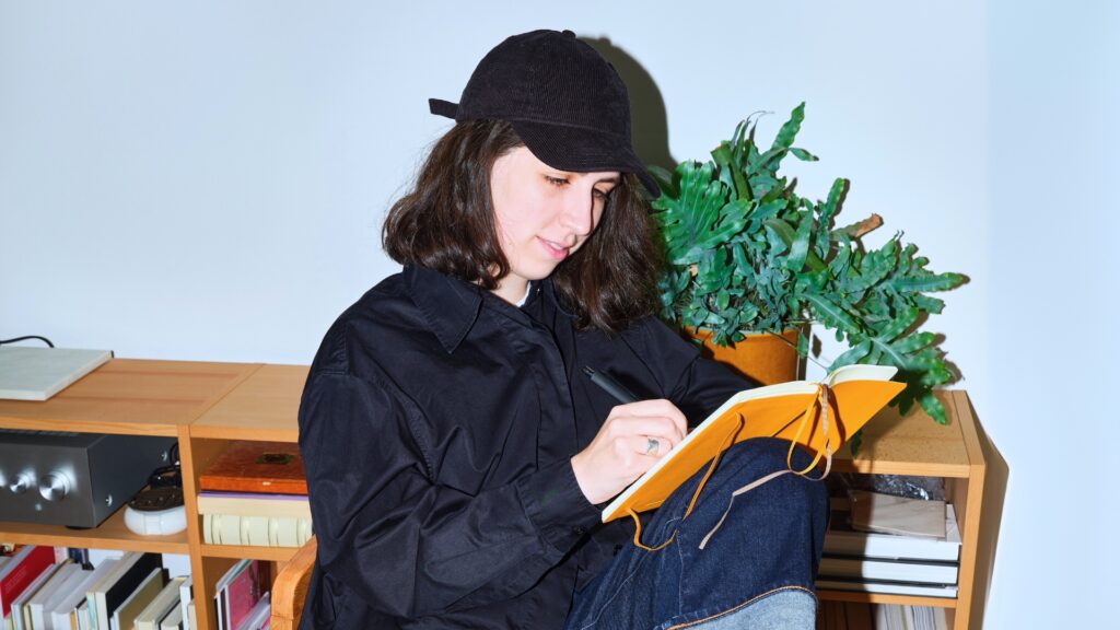 I ett mysigt rum fyllt med böcker och en växt skriver en person med svart keps anteckningar i en orange anteckningsbok, kanske fångar insikter från sina studier på yrkeshögskola.