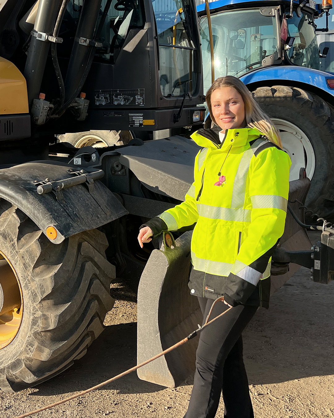 En platschef i en knallgul jacka står självsäkert bredvid entreprenadmaskiner och håller skickligt i en kabel utomhus.