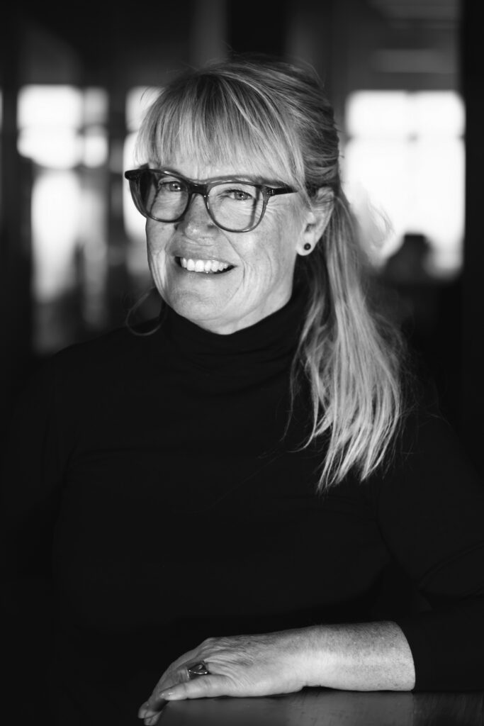 Leende kvinna med glasögon, klädd i en svart ponyhals och hästsvans, i en mjukt upplyst inomhusmiljö.