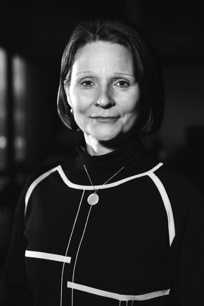 En kvinna med kort hår, iklädd mönstrad topp och halsband, står inomhus och tittar på kameran. Svartvit.