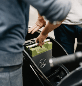 Två personer placerar ett grönt batteri i förvaringsfacket på en elscooter, vilket symboliserar hållbar affärsutveckling inom gårdsförsäljning och en övergång till mer hållbara affärsmetoder.