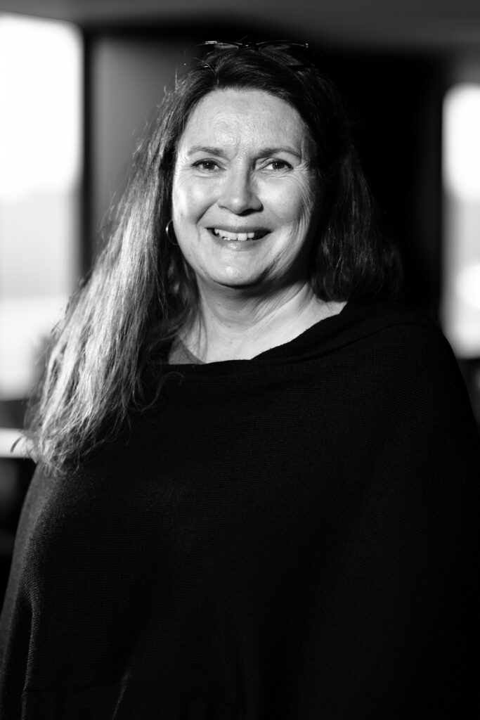 Leende kvinna med långt hår, klädd i en mörk topp, i ett mjukt upplyst rum. Svartvitt fotografi.
