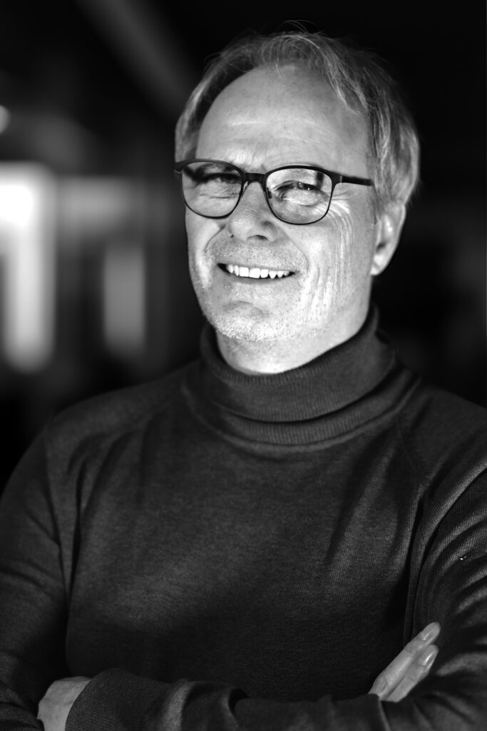 Leende man i glasögon och en turtleneck, armarna i kors, svartvitt foto.