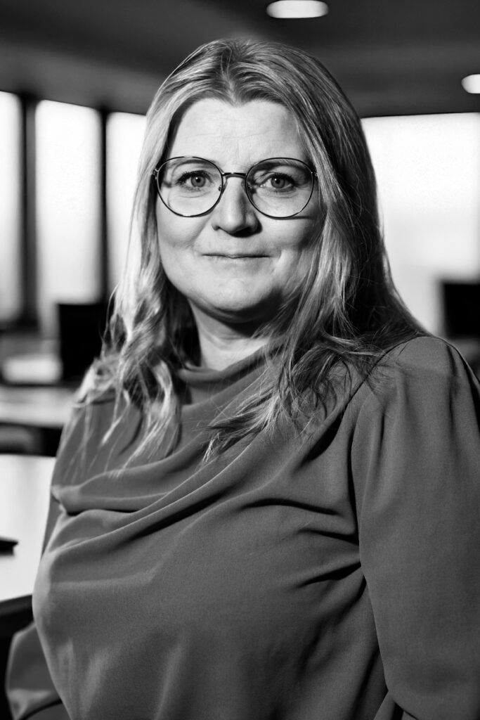 Svartvitt foto av en person med glasögon, långt hår och ett avslappnat uttryck inomhus.