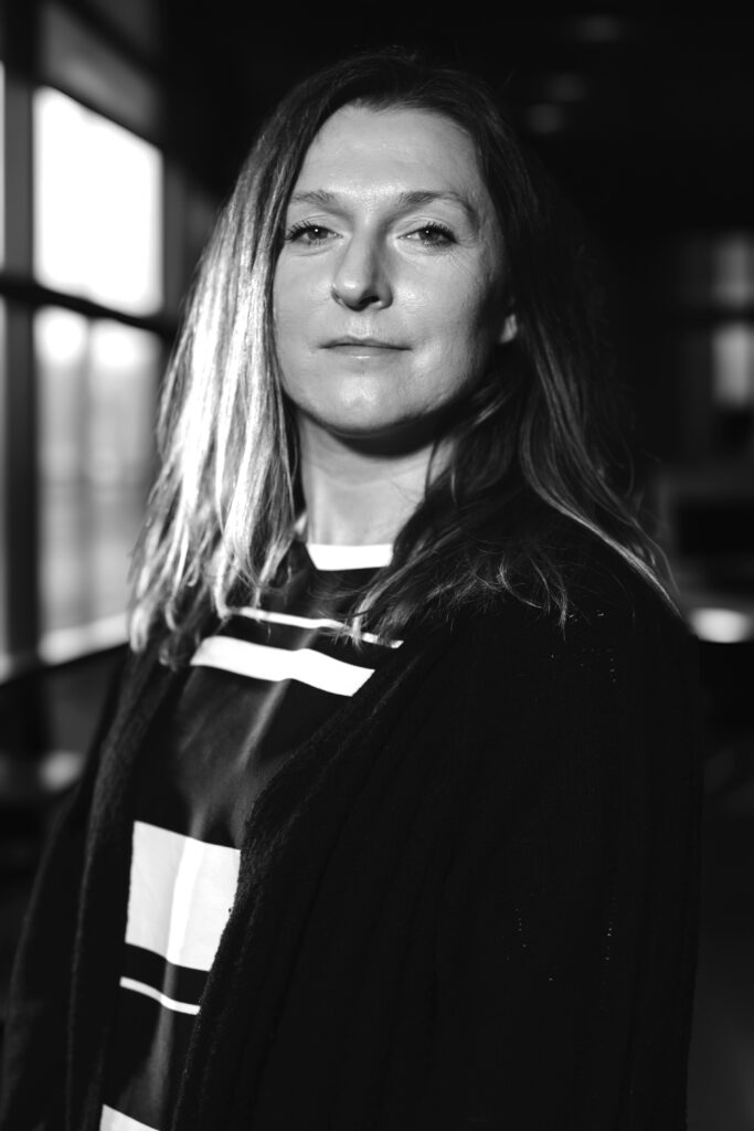 Svartvitt foto av en kvinna med långt hår, stående inomhus, mjukt upplyst från sidan.