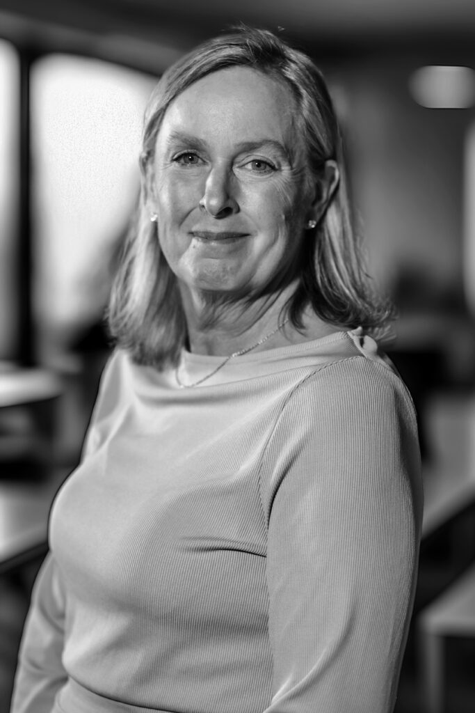 Leende kvinna i en ljus topp sitter inomhus och tittar på kameran med en suddig bakgrund. Svartvit.