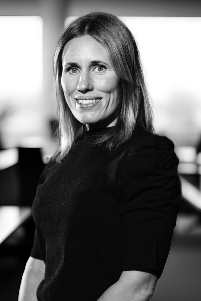 Leende kvinna med axellångt hår klädd i en svart topp, stående i en mjukt upplyst inomhusmiljö. Svartvitt foto.