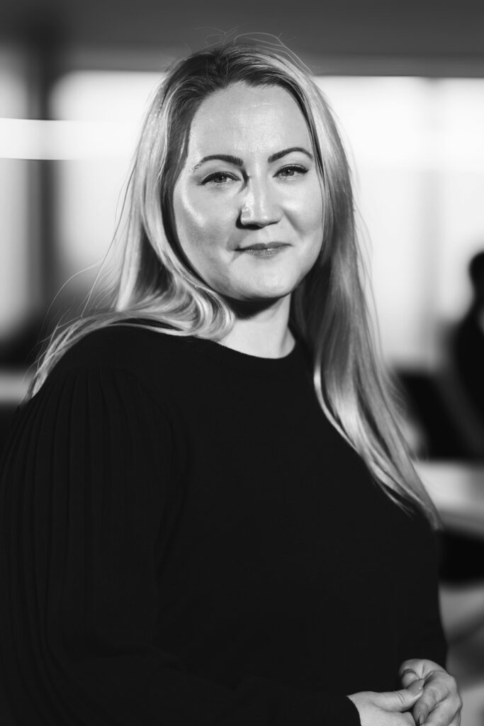 Kvinna med långt hår ler mjukt, klädd i en mörk topp, i ett svagt upplyst rum. Svartvitt foto.
