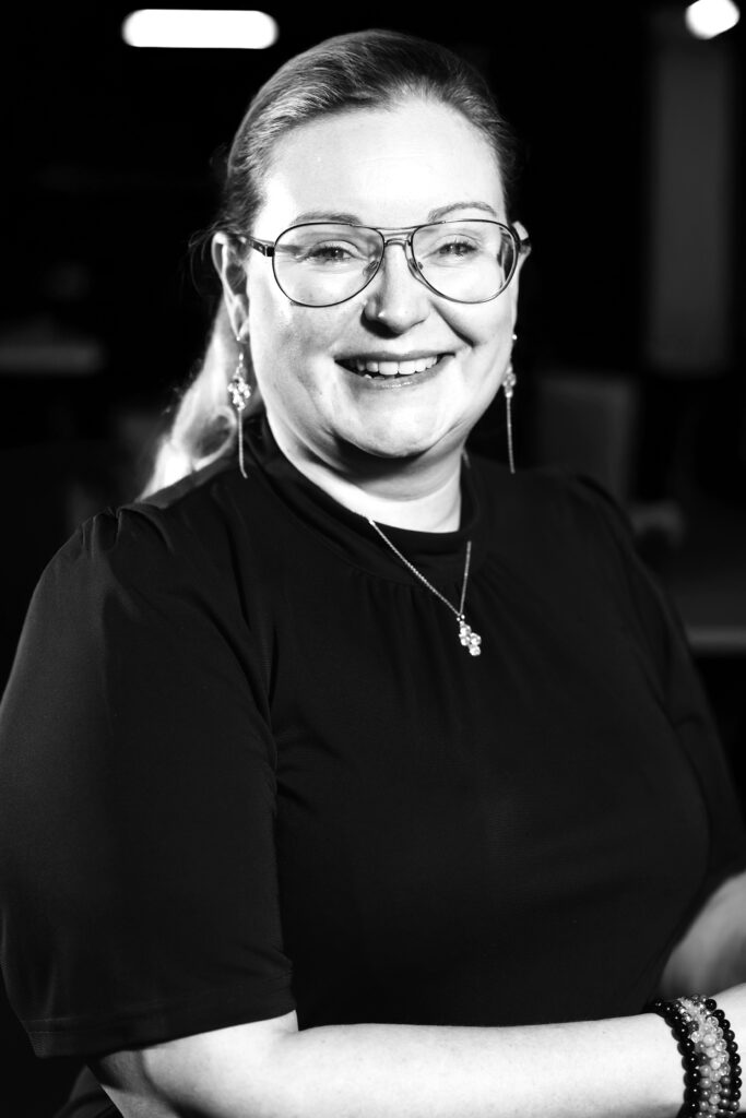 Person som bär glasögon och ett korshalsband och ler under inomhusbelysning. Svartvitt foto.
