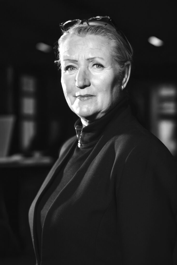 Äldre kvinna i en svart jacka med glasögon på huvudet, stående i ett svagt upplyst rum. Svartvit bild.