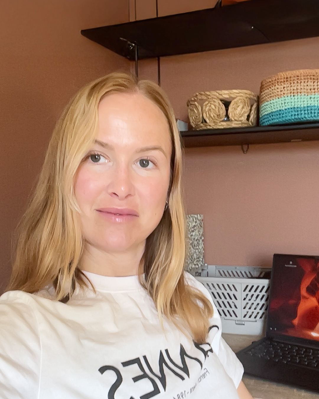 En blondhårig person, ler i ett rum med en bärbar dator och hyllor i bakgrunden, förkroppsligar andan i en byggnadsingenjör inom Energi och ROT, som harmoniserar teknik med hållbar innovation.